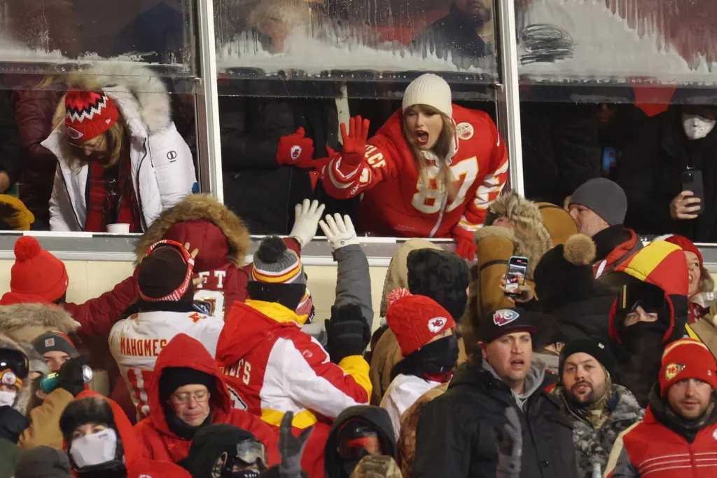 Taylor Swift in a Chiefs jersey with a white beanie, waving to fans at the AFC Wild Card Playoffs.