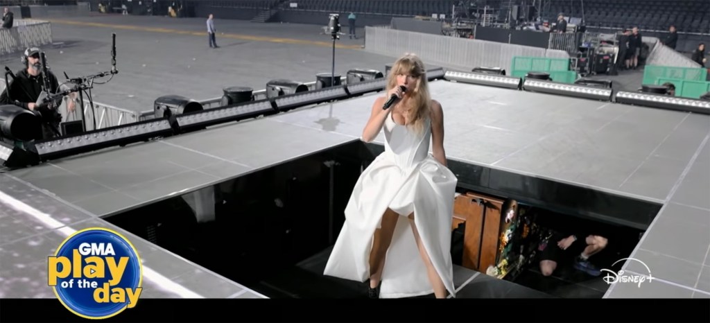 Taylor Swift in a white dress and holding a microphone while performing on a stage.