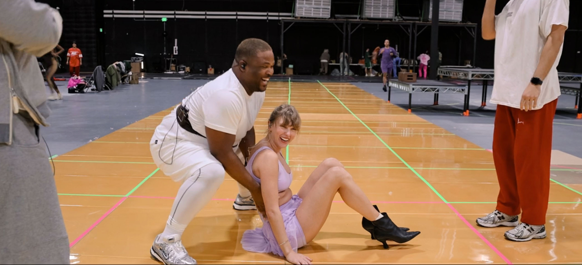 Taylor Swift in a light purple outfit, laughing on the floor, while a male dancer in white leans over her.
