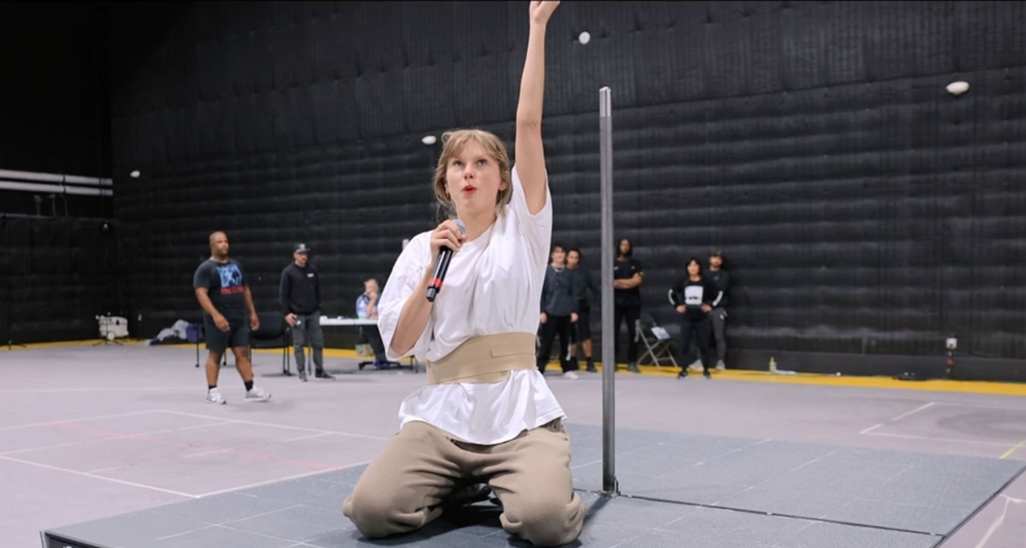 Taylor Swift rehearsing on her knees, holding a microphone, with her right arm raised in a large empty studio with crew members in the background.