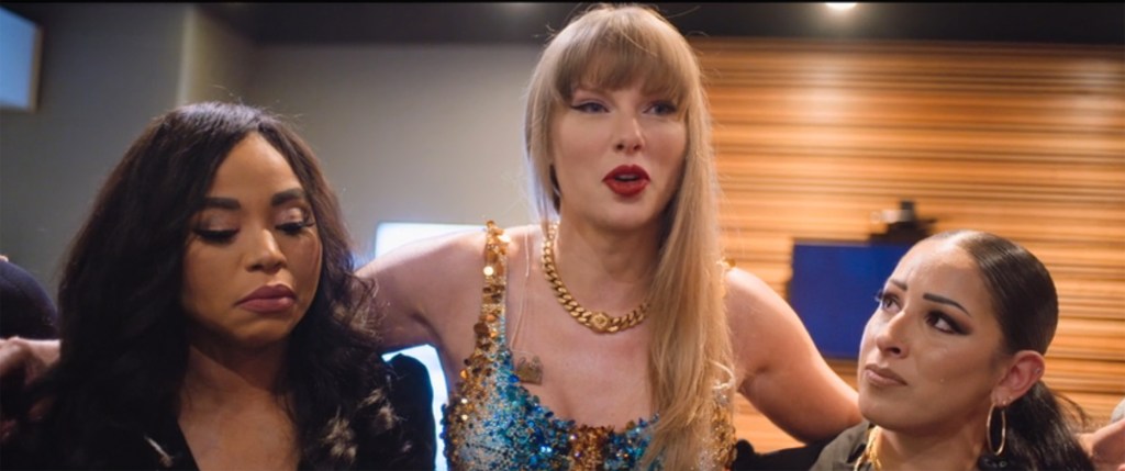 Taylor Swift on stage in a sparkly dress, talking to two people, as seen from the