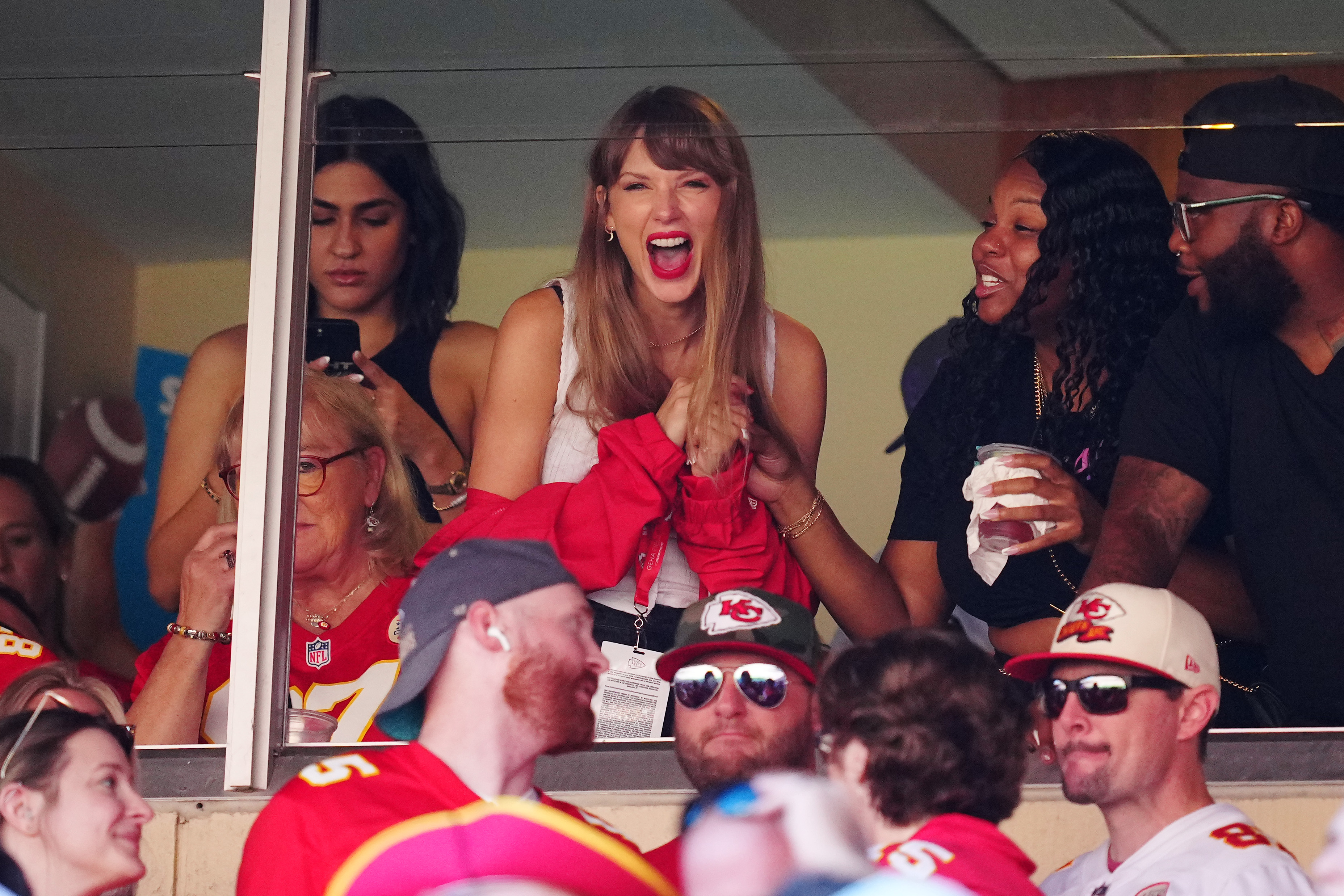 Taylor Swift, wearing a white top and red jacket, cheers excitedly from a stadium box at the Chiefs vs. Bears game.