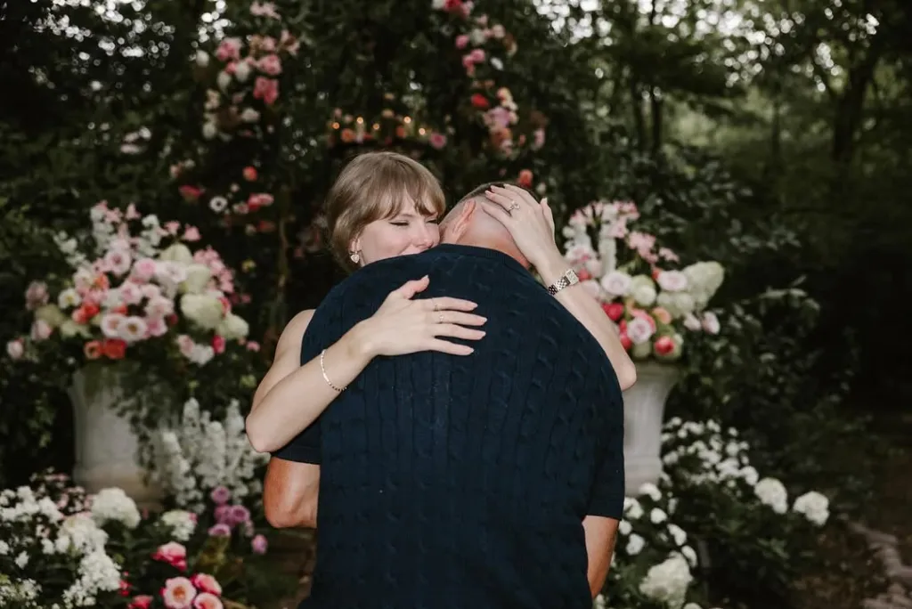 Taylor Swift hugging Travis Kelce, with engagement rings visible on both their hands, surrounded by flowers.