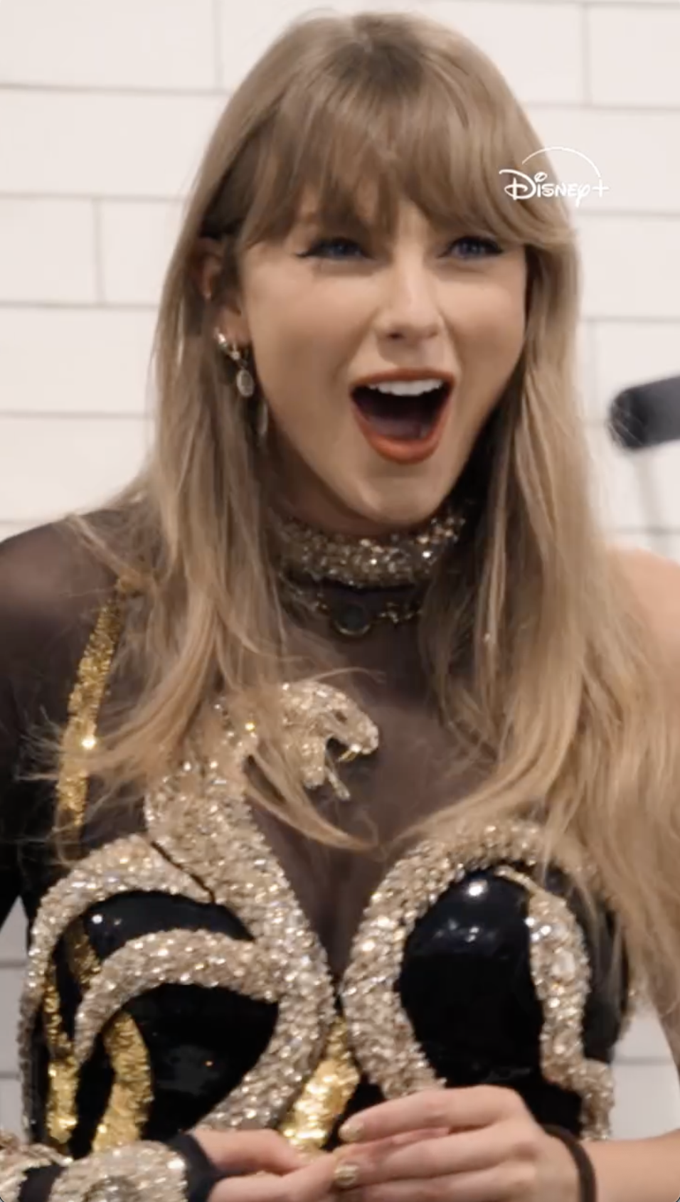 Taylor Swift in a black and gold sequined outfit with a snake motif on the bodice, mouth open in excitement.