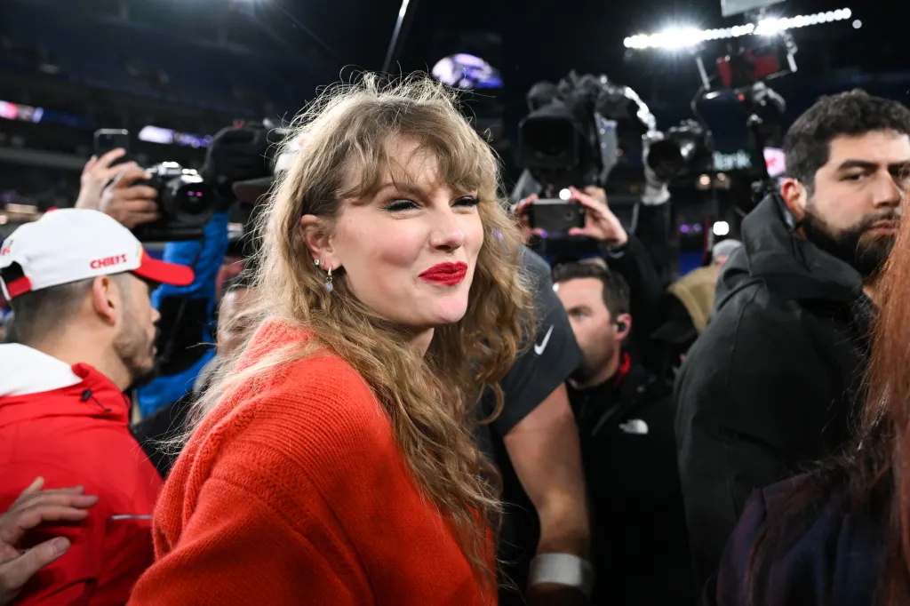 Taylor Swift in a red sweater, with a crowd and cameras behind her, during the AFC Championship.