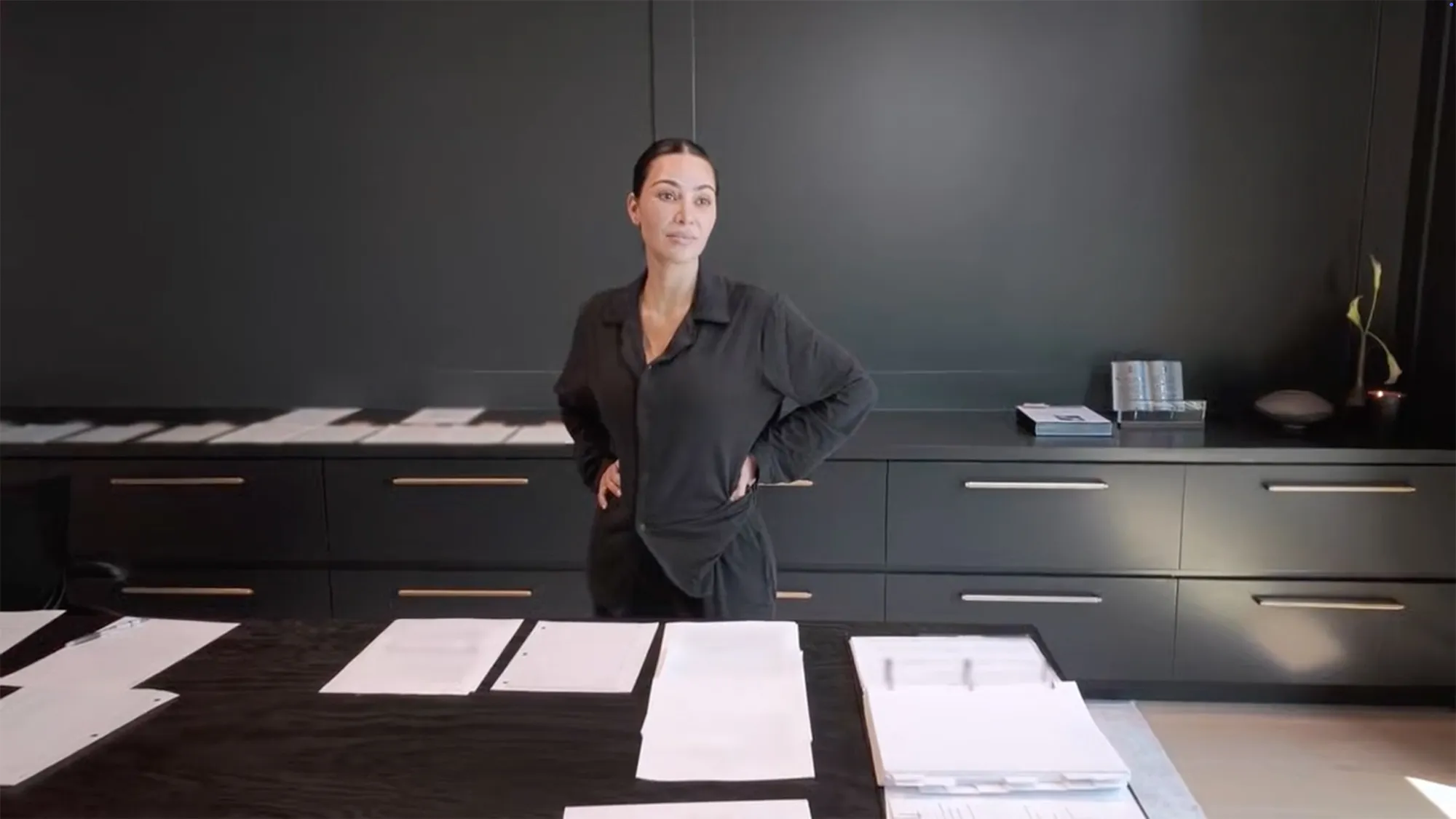 Kim Kardashian, dressed in black, standing in a dark room with papers on a table, expressing discomfort during her bar exam studies.