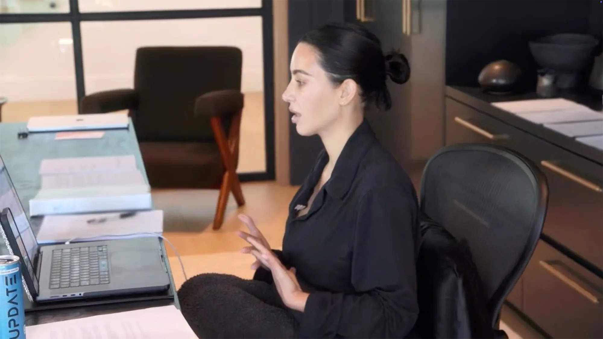 Kim Kardashian speaking while sitting at a desk with a laptop and papers.