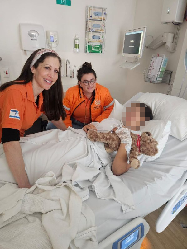 Two women in orange shirts visiting a patient in a hospital bed, the patient's face is blurred, but their bandaged hand is visible holding a teddy bear.