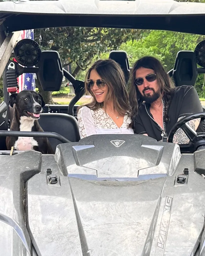 Elizabeth Hurley and Billy Ray Cyrus in a utility vehicle with a dog.
