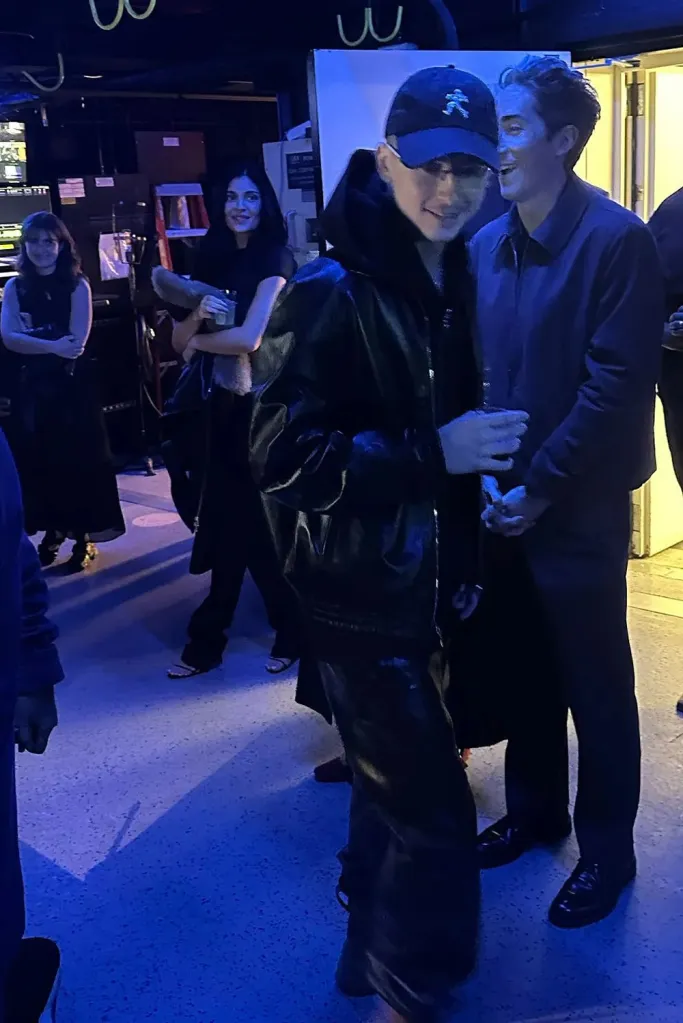 Timothée Chalamet wearing a blue cap and black jacket, with Kylie Jenner , in a black dress, in the background, at the NYFF Marty Supreme premiere.