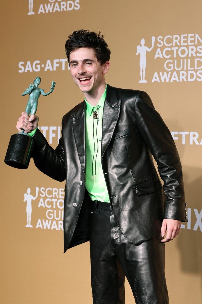 Timothée Chalamet, in a black leather blazer, green shirt and black leather pants, smiling and holding his SAG award for Outstanding Performance by a Male Actor.