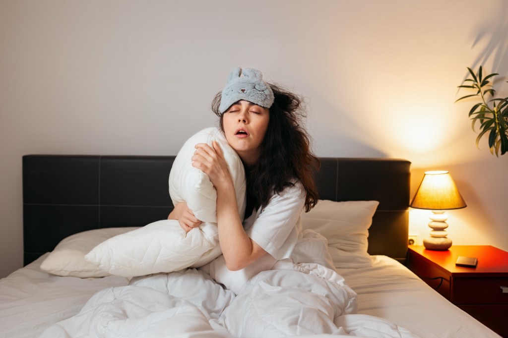 Tired woman with a sleep mask sitting in bed hugging a pillow, unable to sleep.