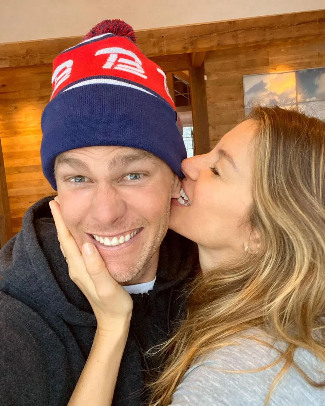 Tom Brady in a Patriots hat with a woman whispering in his ear.