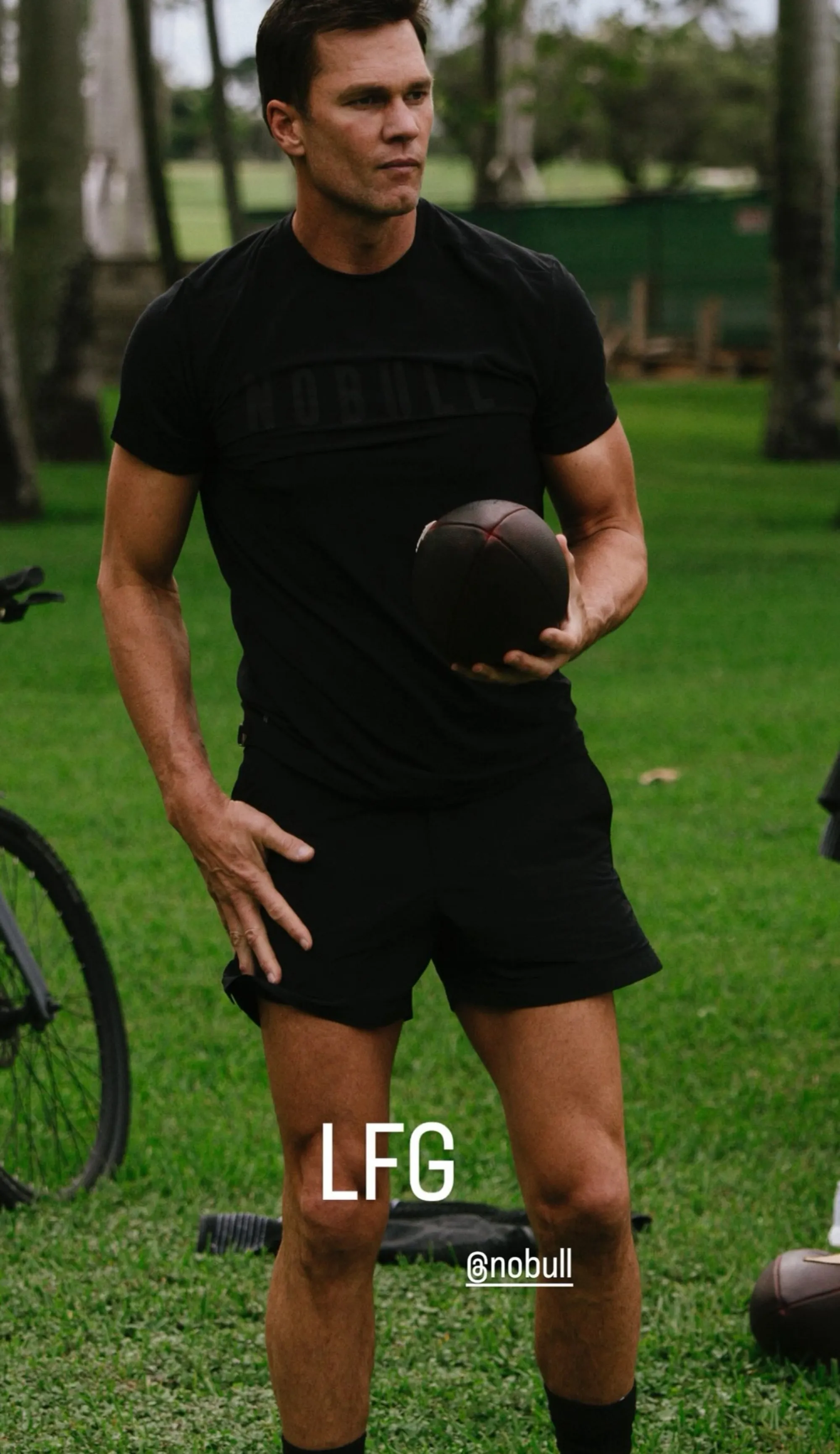 Tom Brady in a black shirt and shorts, holding a football.