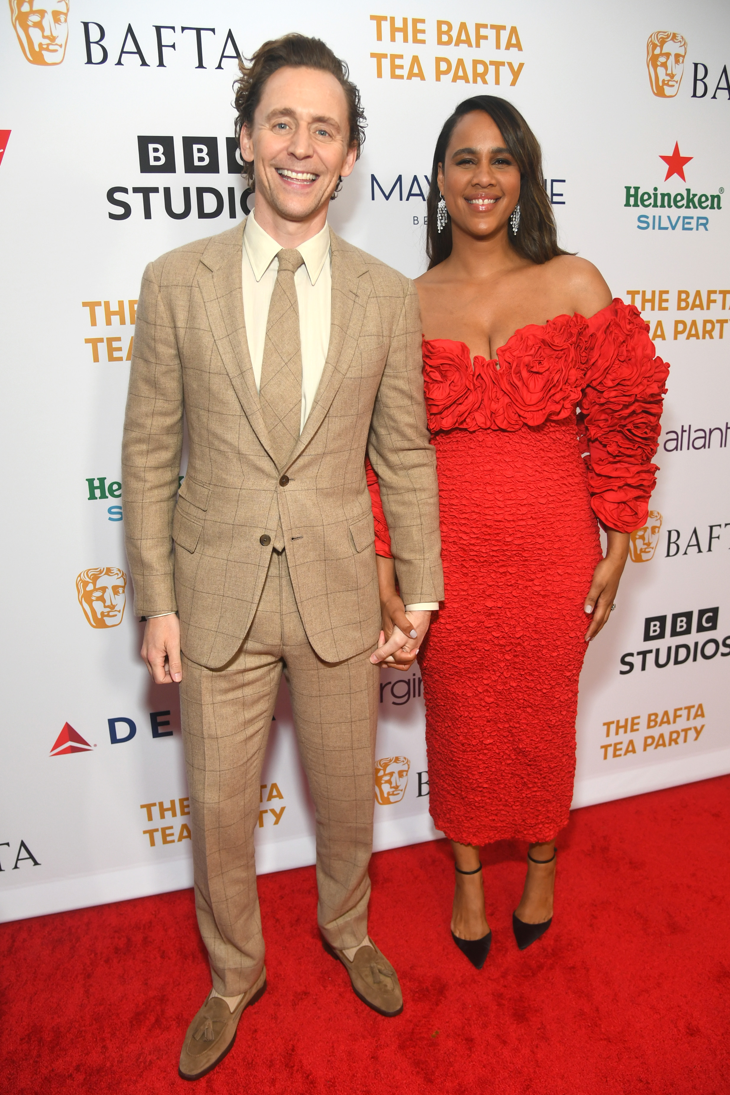 Tom Hiddleston in a brown plaid suit and Zawe Ashton in a red ruffled dress hold hands at the BAFTA Tea Party.
