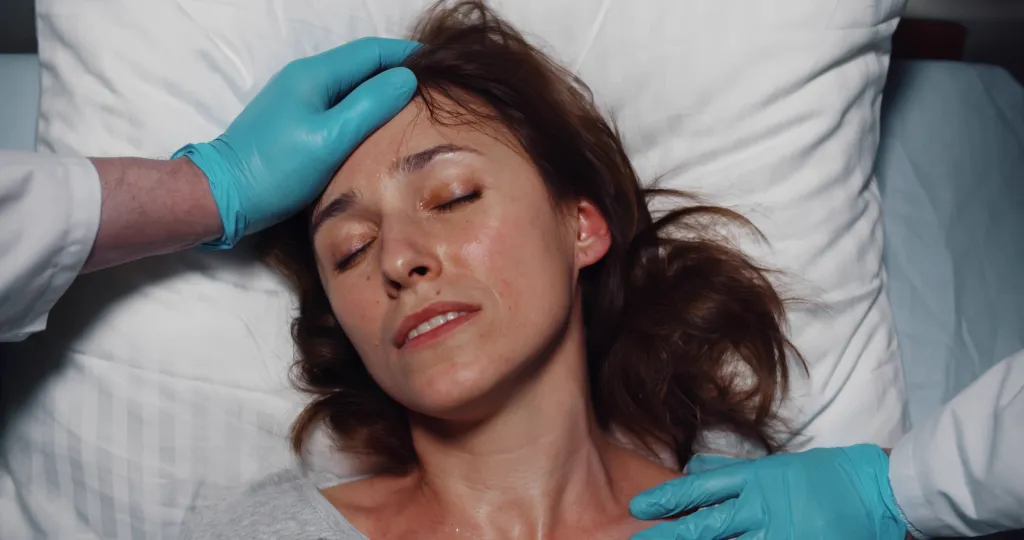 Top view of female patient with high fever lying unconscious in hospital bed, with doctor checking her temperature.