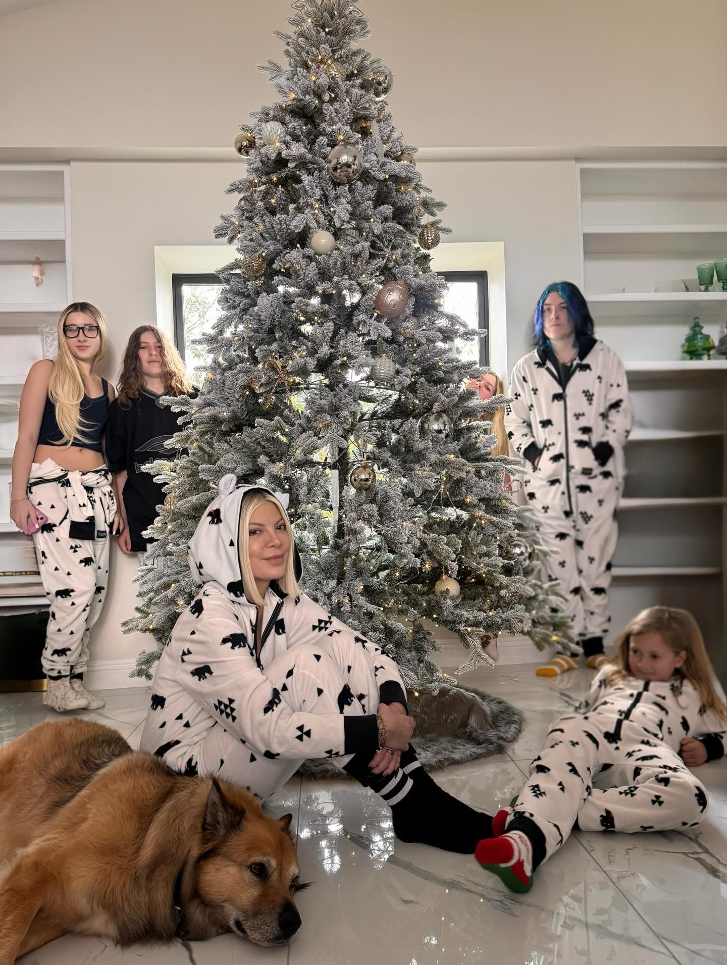 Tori Spelling and her children in matching bear pajamas next to a flocked Christmas tree.