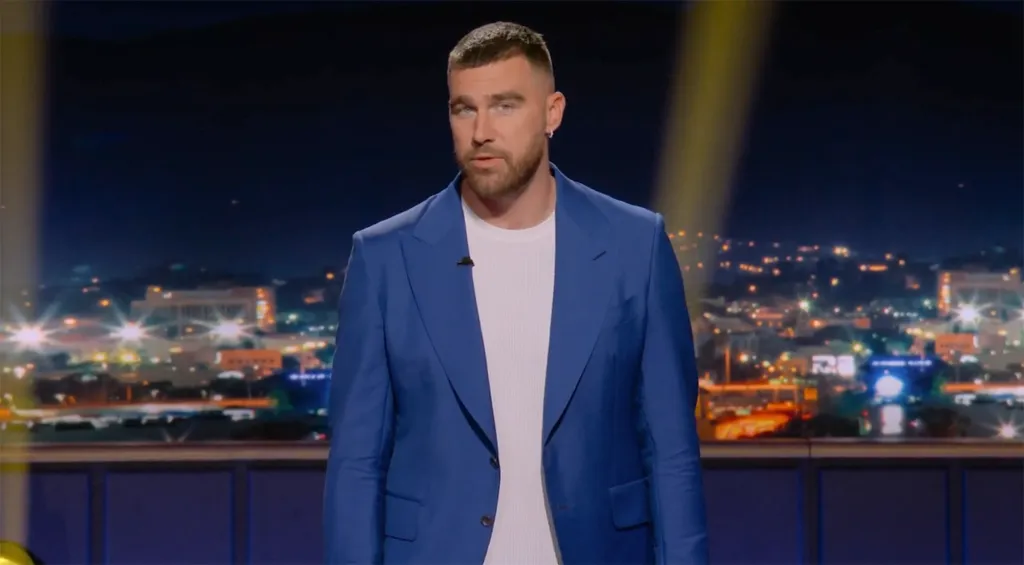 Travis Kelce on a game show, wearing a blue blazer and white t-shirt, with a city skyline at night in the background.