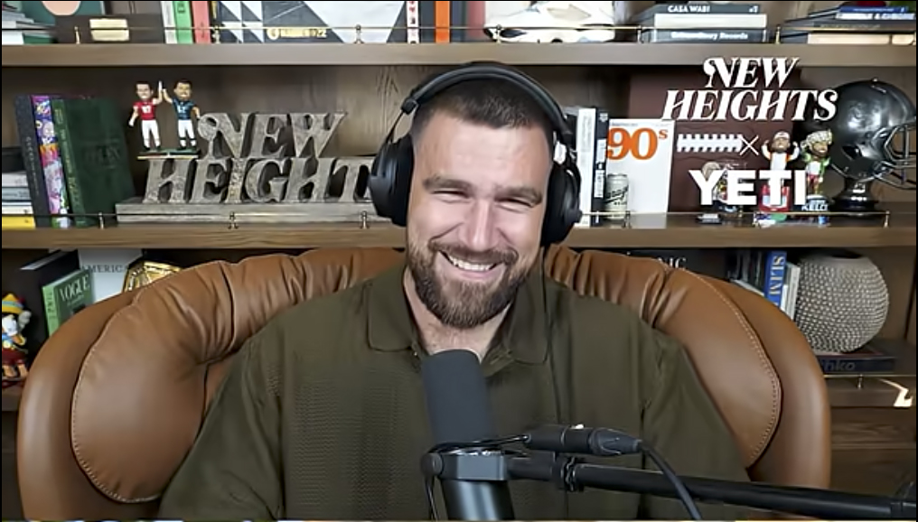 Travis Kelce, wearing headphones, laughs into a microphone during the