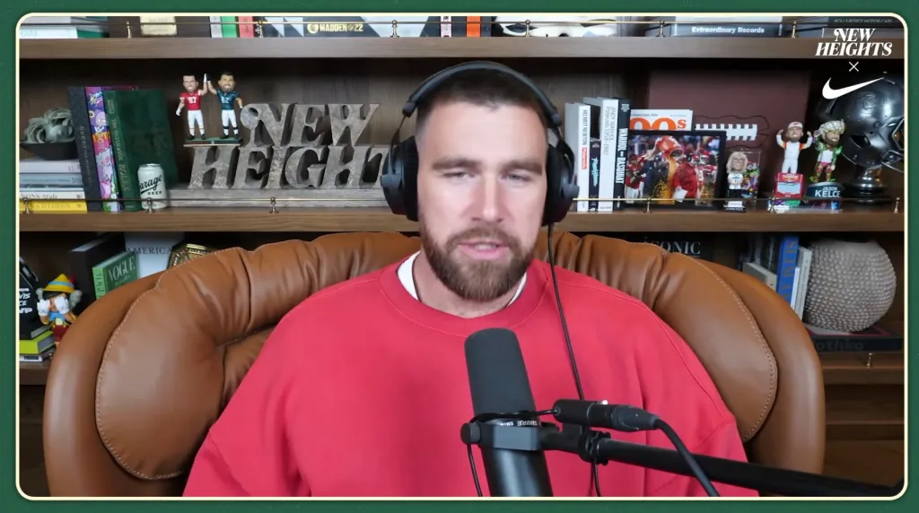 Travis Kelce wearing headphones and a red sweatshirt, speaking into a microphone, with a shelf of books and sports memorabilia in the background.