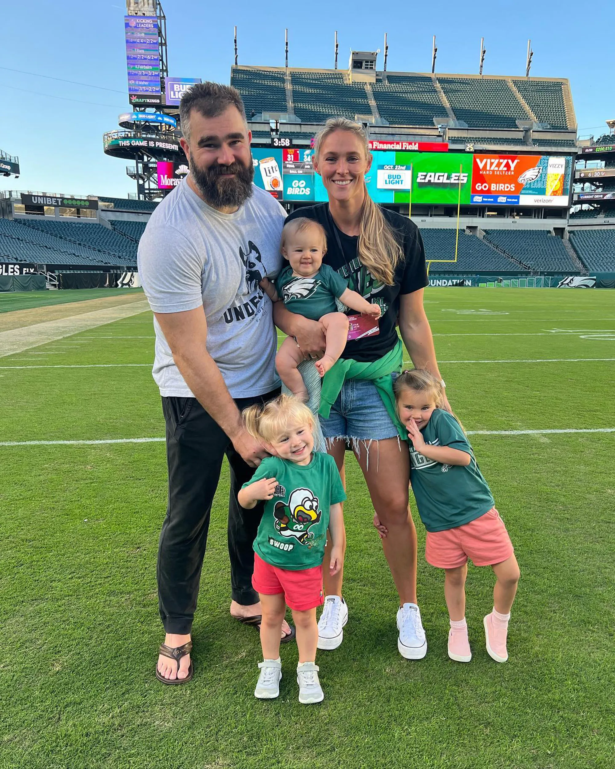 Kylie Kelce, Jason Kelce, and their three daughters pose on a football field in front of a stadium.