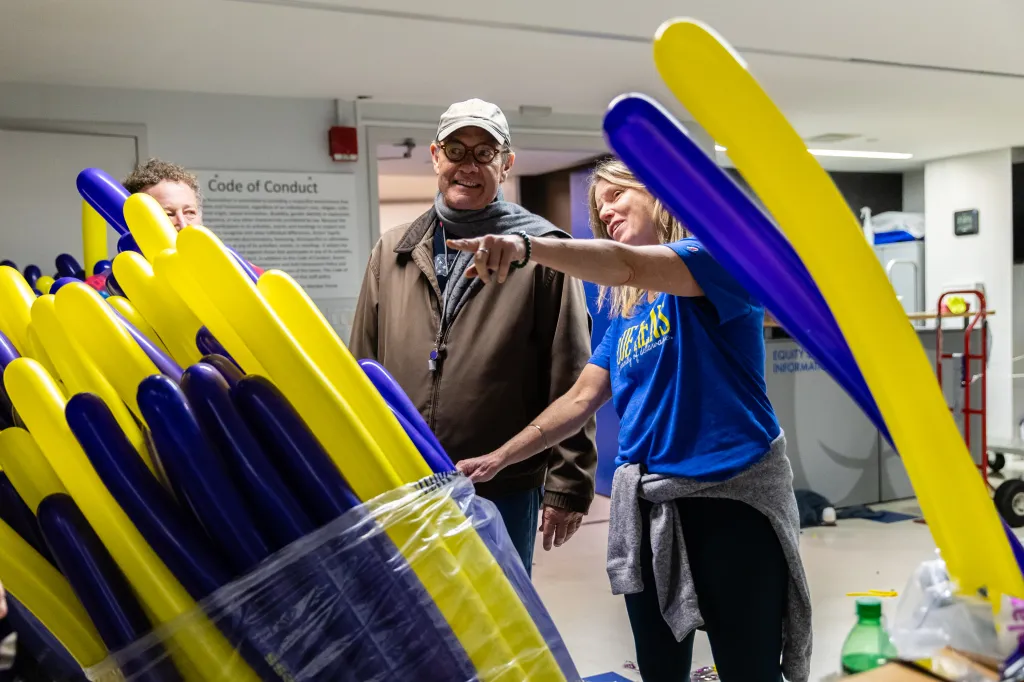 Three people, one with curly hair, one man wearing a hat, and a woman wearing a blue shirt, stand among boxes filled with yellow and purple confetti sticks.