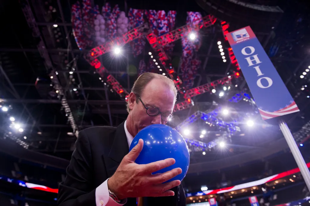 Treb Heining blowing up a blue balloon on the convention floor with a sign for 