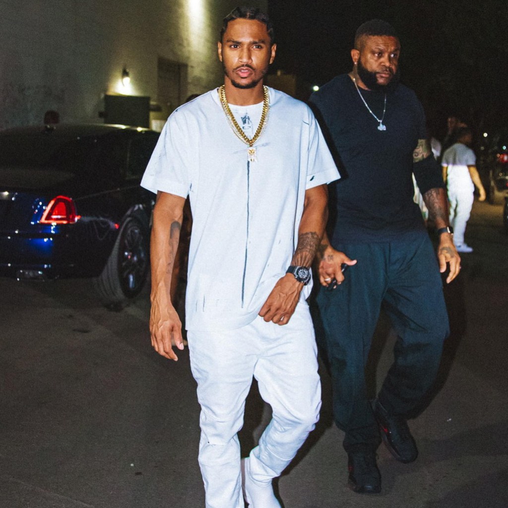 Trey Songz walks with a man in a dark background.