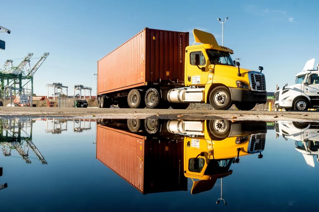 A yellow truck with a red shipping container parked at a port is reflected in a puddle.