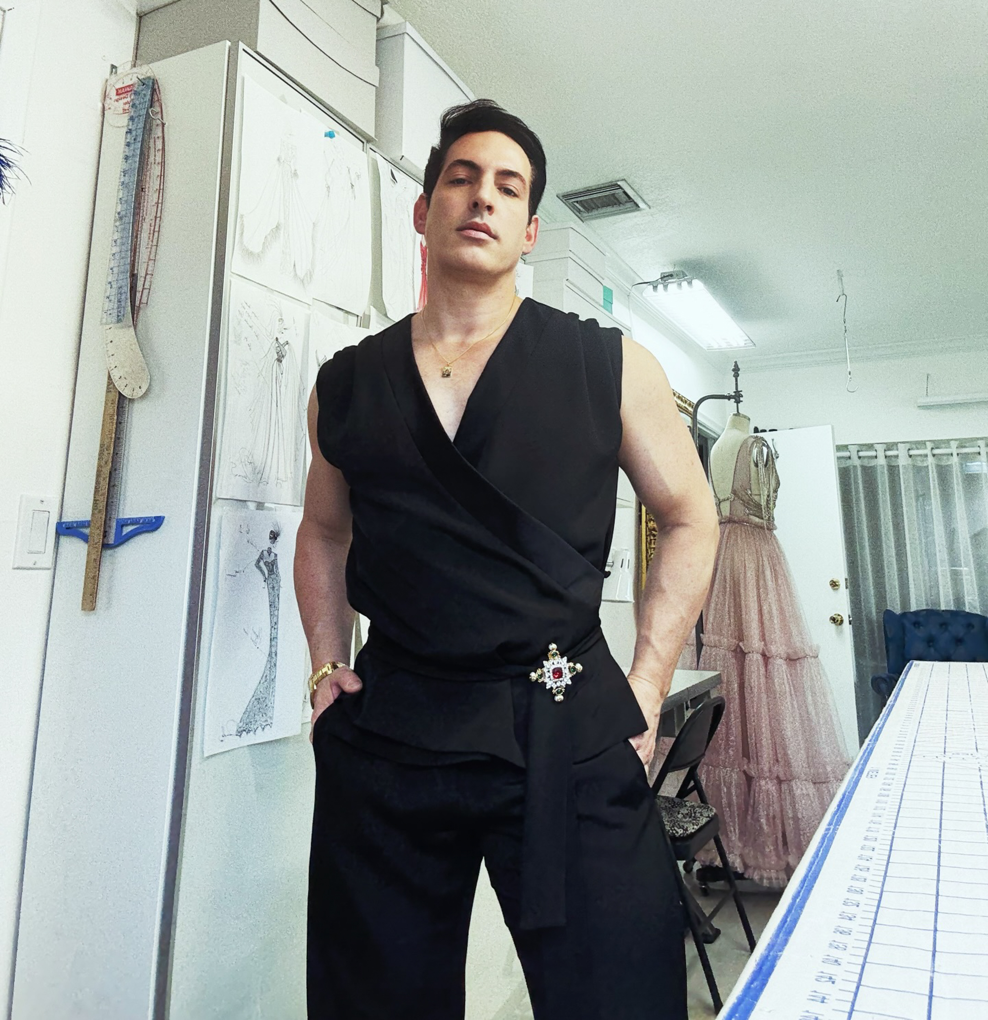 Fashion designer Oscar G. Lopez in a black top with an ornate broach and black pants in his studio.