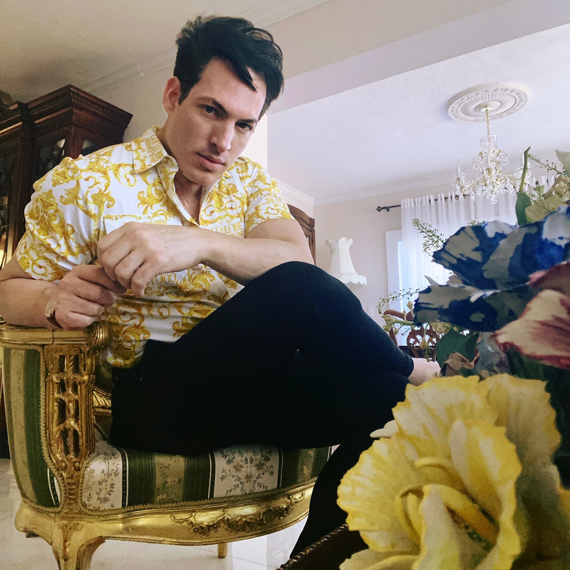 Oscar G. Lopez pictured sitting on a gold and green upholstered chair, wearing a white short-sleeved collared shirt with yellow designs and black pants.