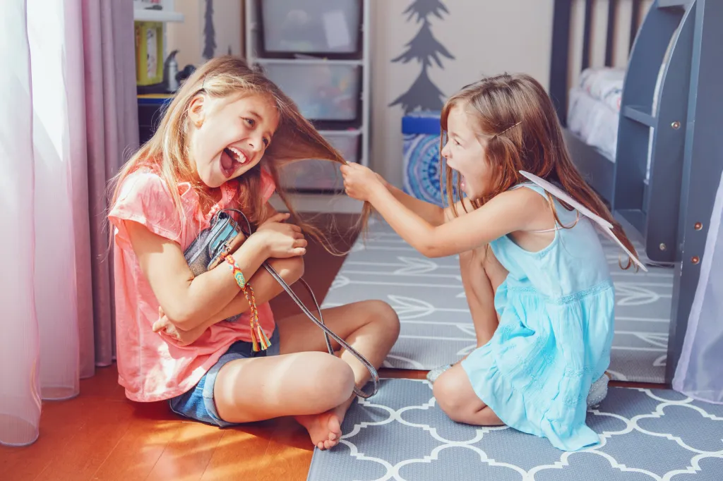 Two young girls, sisters, fighting with one pulling the other's hair, in a brightly lit room.