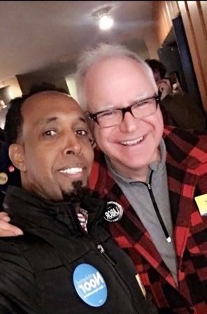 Two men, one in a black jacket and the other in a red and black plaid jacket, smiling.