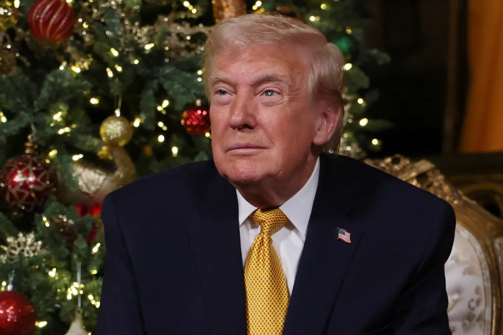 U.S. President Donald Trump in a suit and yellow tie with an American flag pin, sitting in front of a decorated Christmas tree.