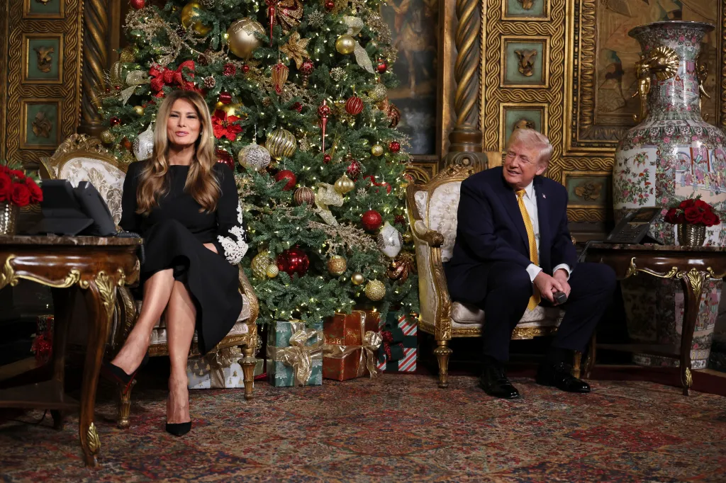Donald and Melania Trump sit in ornate chairs in front of a Christmas tree decorated with red and gold ornaments, with presents at its base.