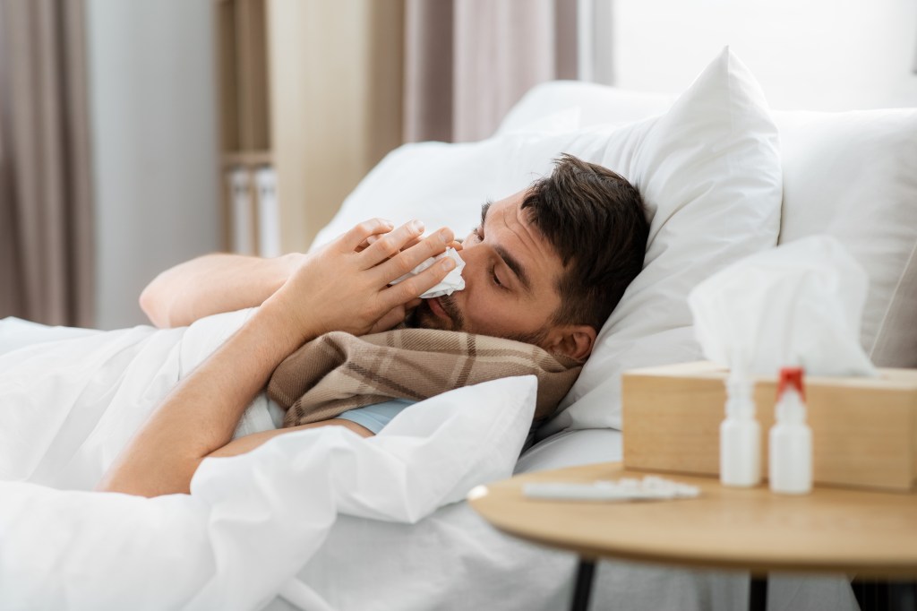 A sick man in bed blowing his nose into a tissue.