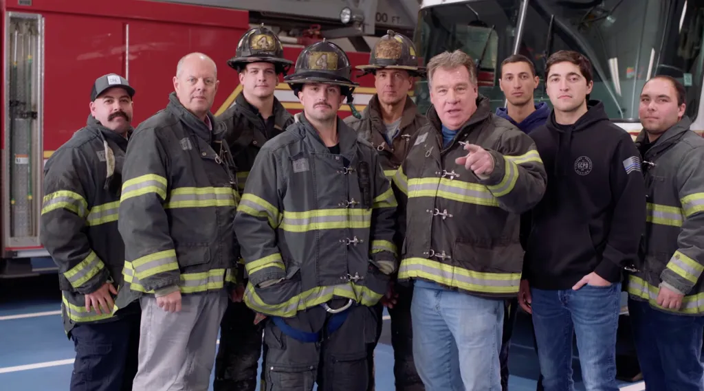A group of firefighters in front of a fire engine, one man pointing.