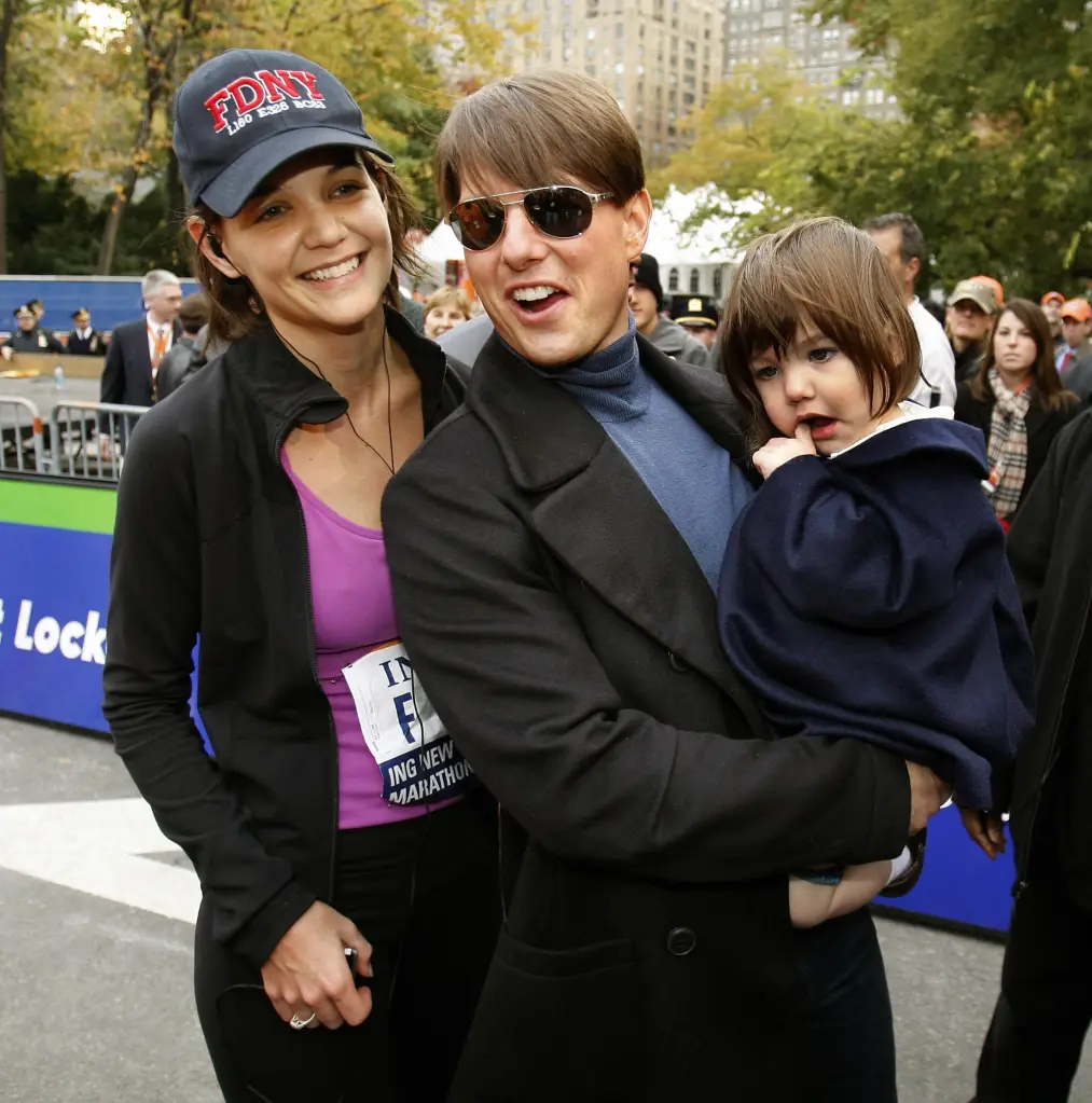 Tom Cruise holding his daughter Suri, standing next to his then-wife Katie Holmes, all smiling at the finish line of the New York City Marathon.