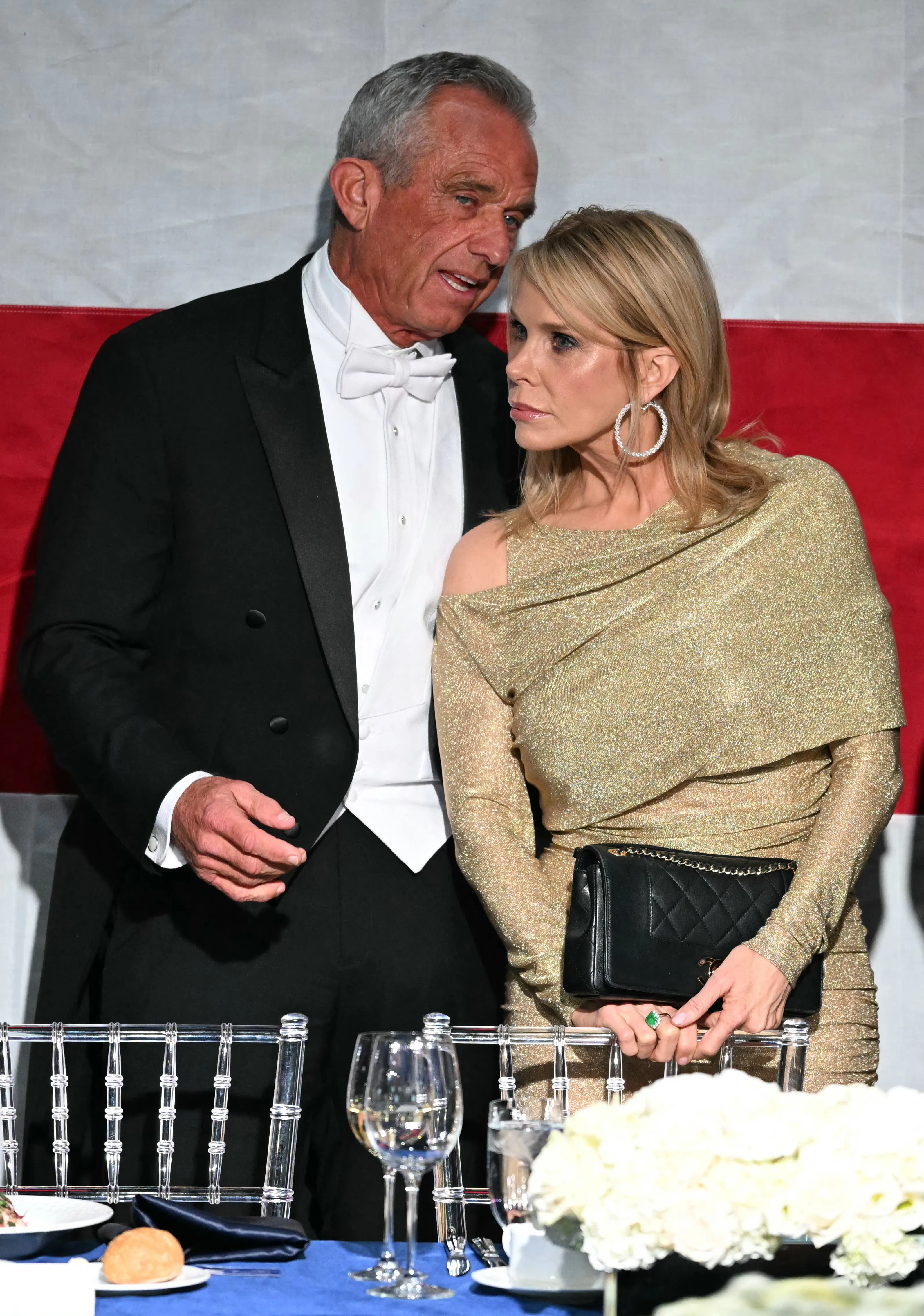 Robert F. Kennedy Jr. in a tuxedo speaking to his wife Cheryl Hines, who is wearing a gold dress and holding a black purse.