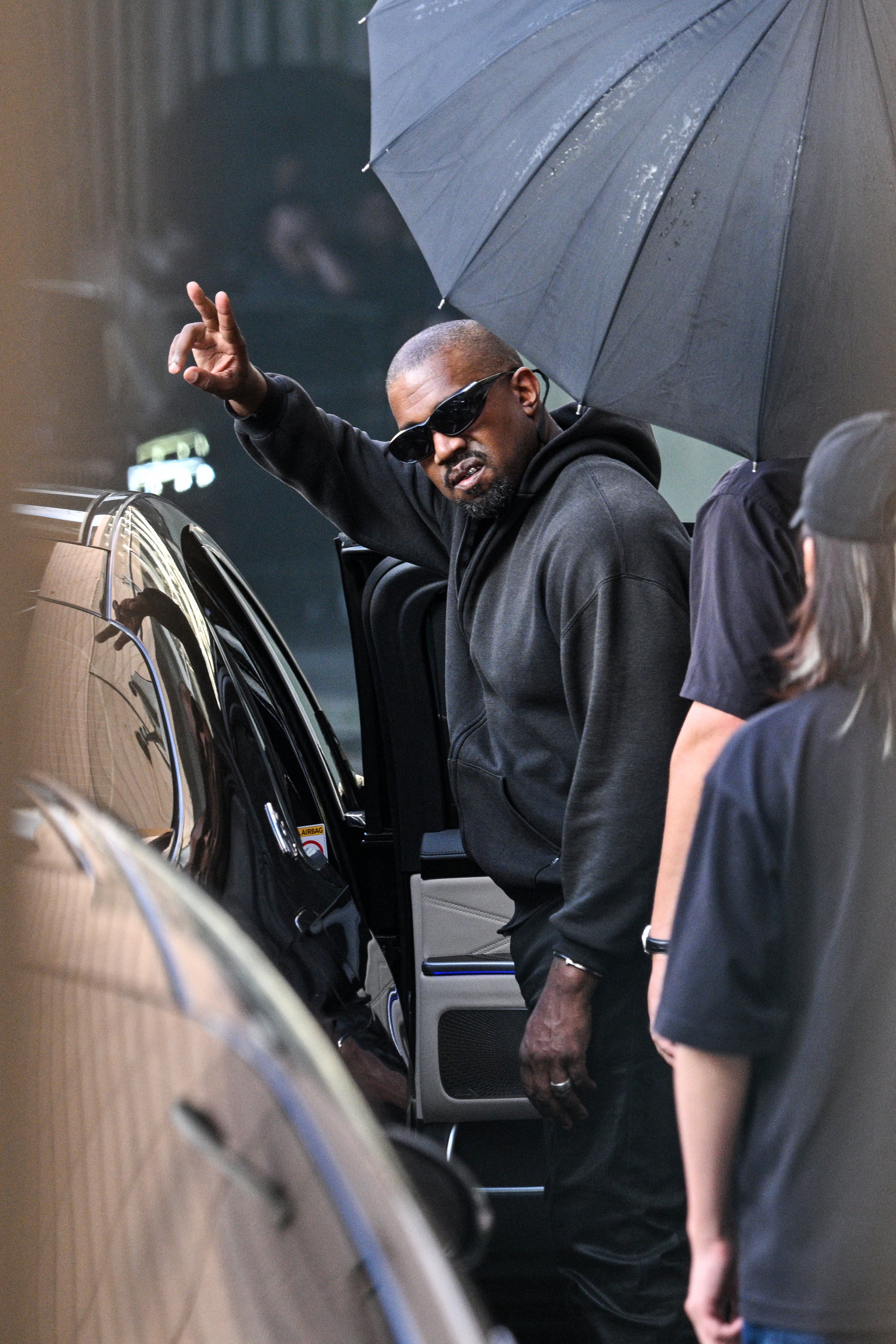 Kanye West wearing sunglasses, a dark hoodie, and grimacing, waves while getting out of a car.
