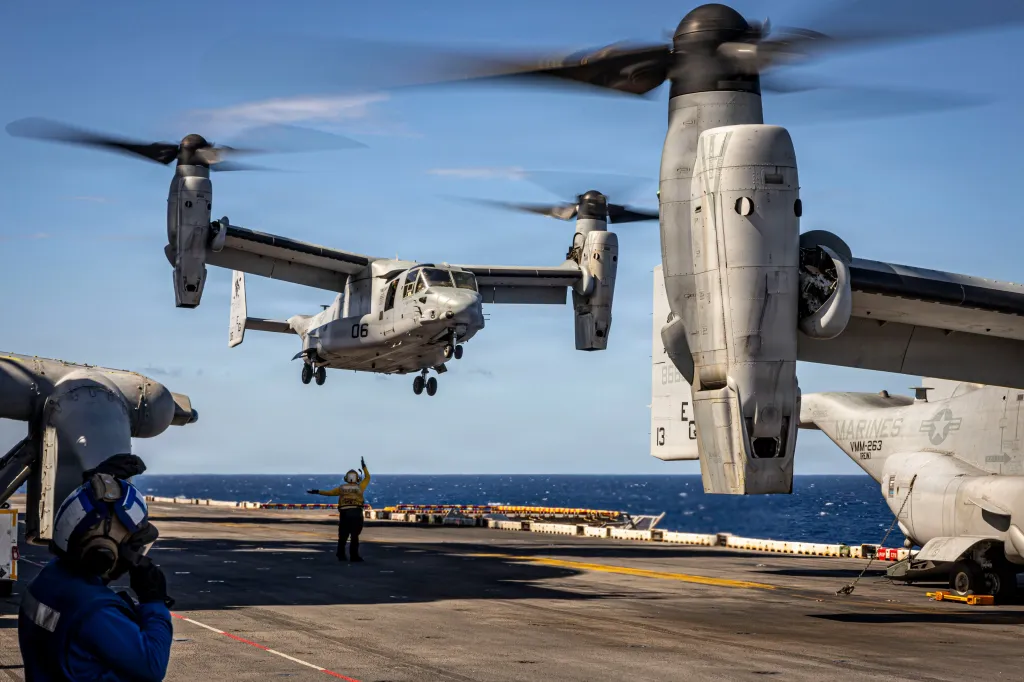 A US Marine Corps MV-22B Osprey tiltrotor aircraft landing on the USS Iwo Jima amphibious assault ship, guided by a sailor, while another Osprey is parked on the deck.