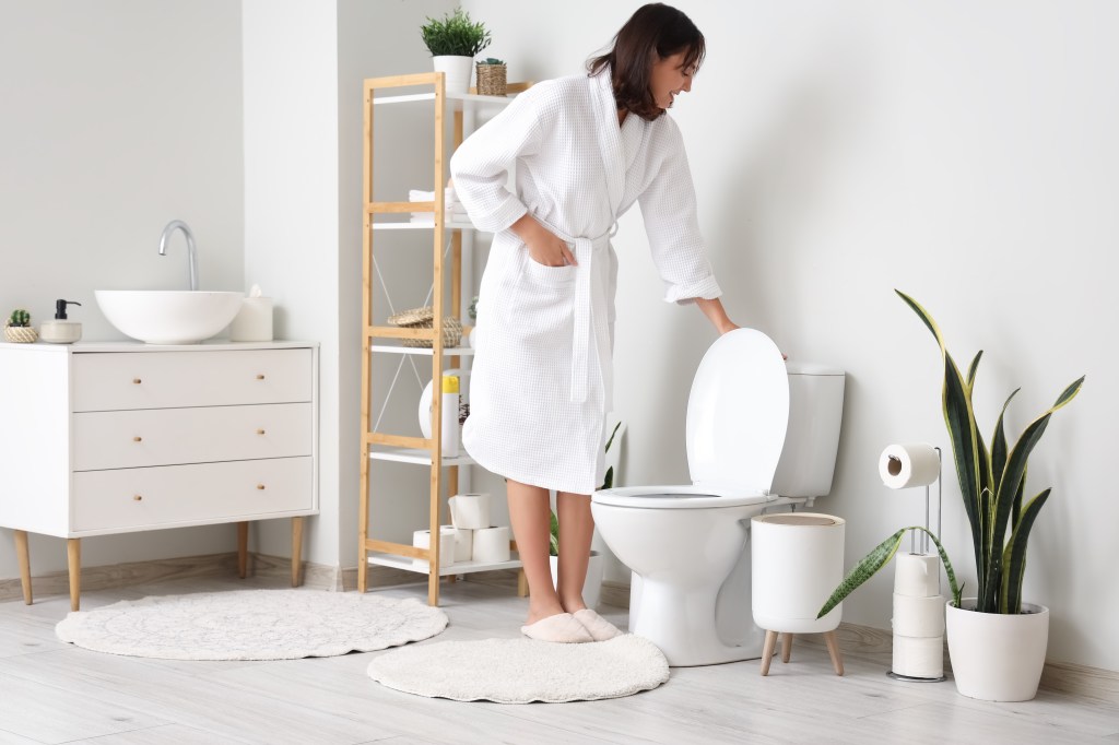 Young woman in a white bathrobe flushing the toilet in a light-colored bathroom.