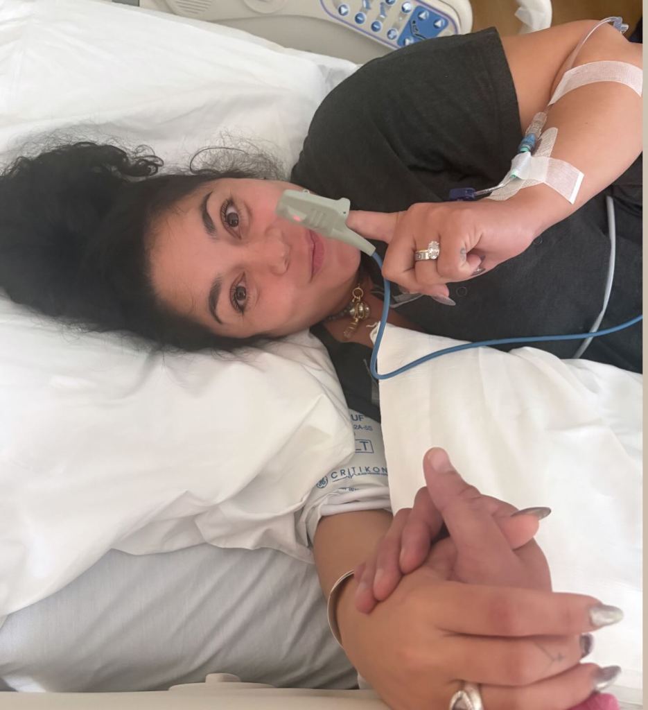 Vanessa Hudgens lying in a hospital bed with an IV drip and a pulse oximeter on her finger, holding hands with someone.