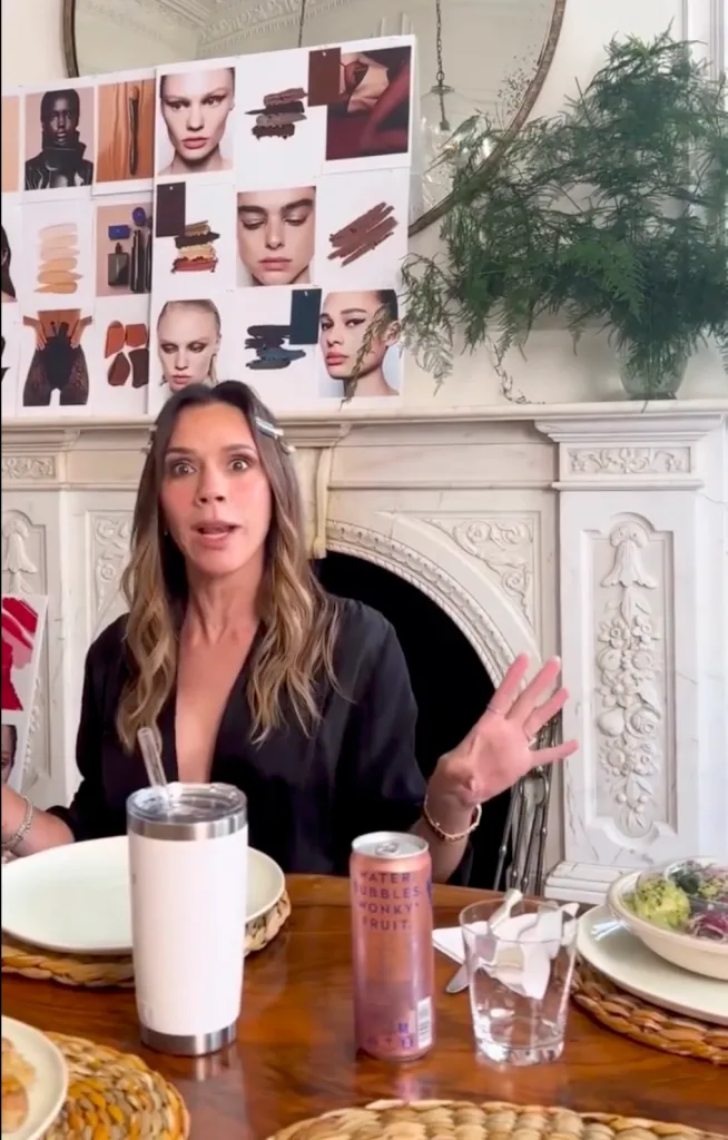 Victoria Beckham sitting at a table with an assortment of makeup inspiration images on a board behind her.
