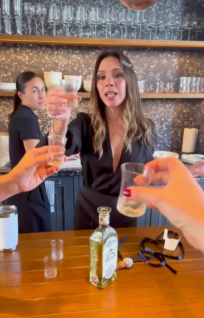 Victoria Beckham toasts with a shot glass of tequila, surrounded by friends also holding shot glasses and a bottle of tequila on a table.