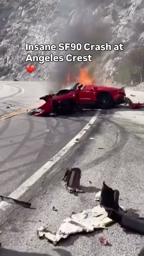 A crashed red Ferrari SF90 on fire on a winding mountain road.