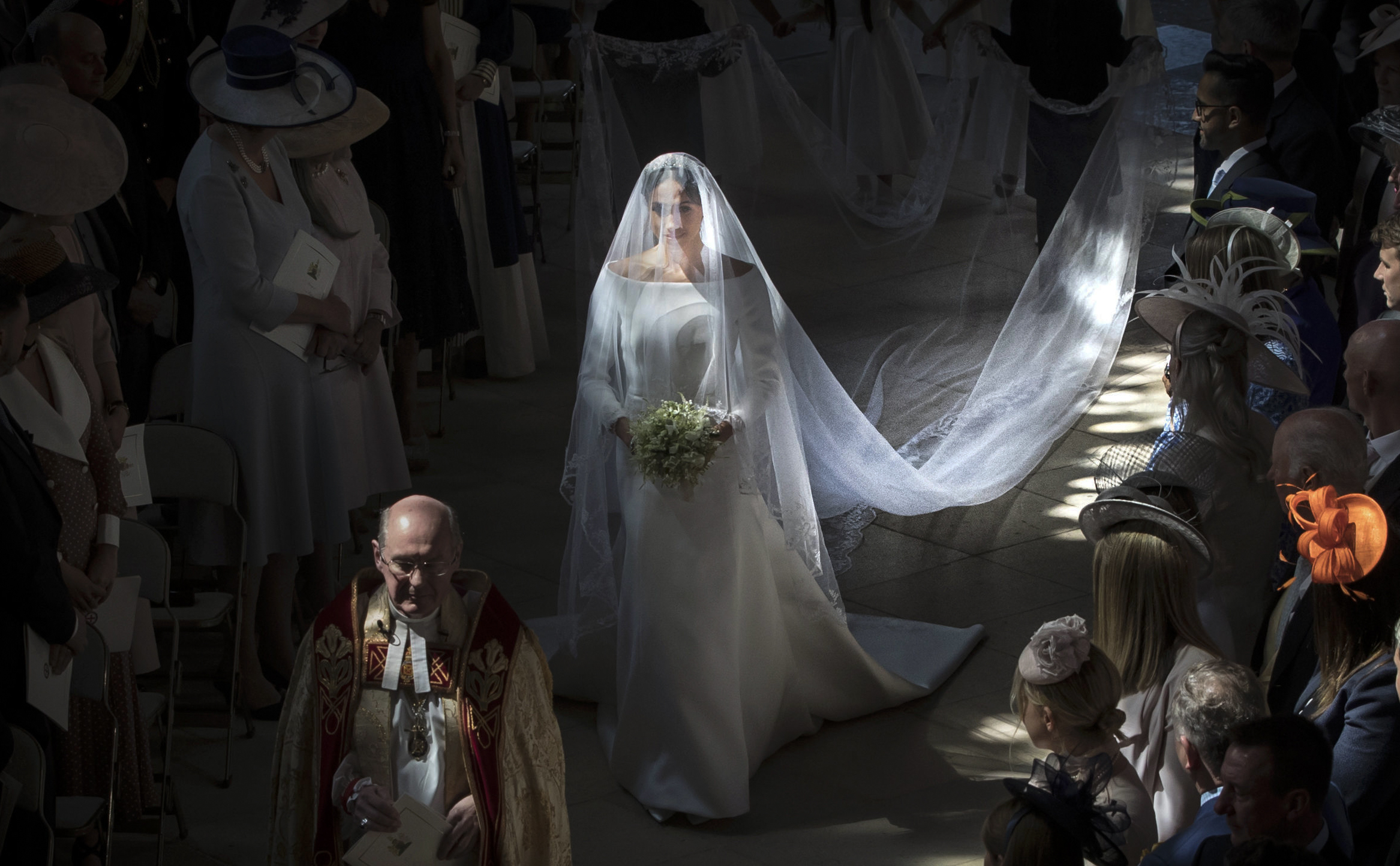 Meghan Markle walks down the aisle in a white wedding dress and long veil, holding a bouquet.