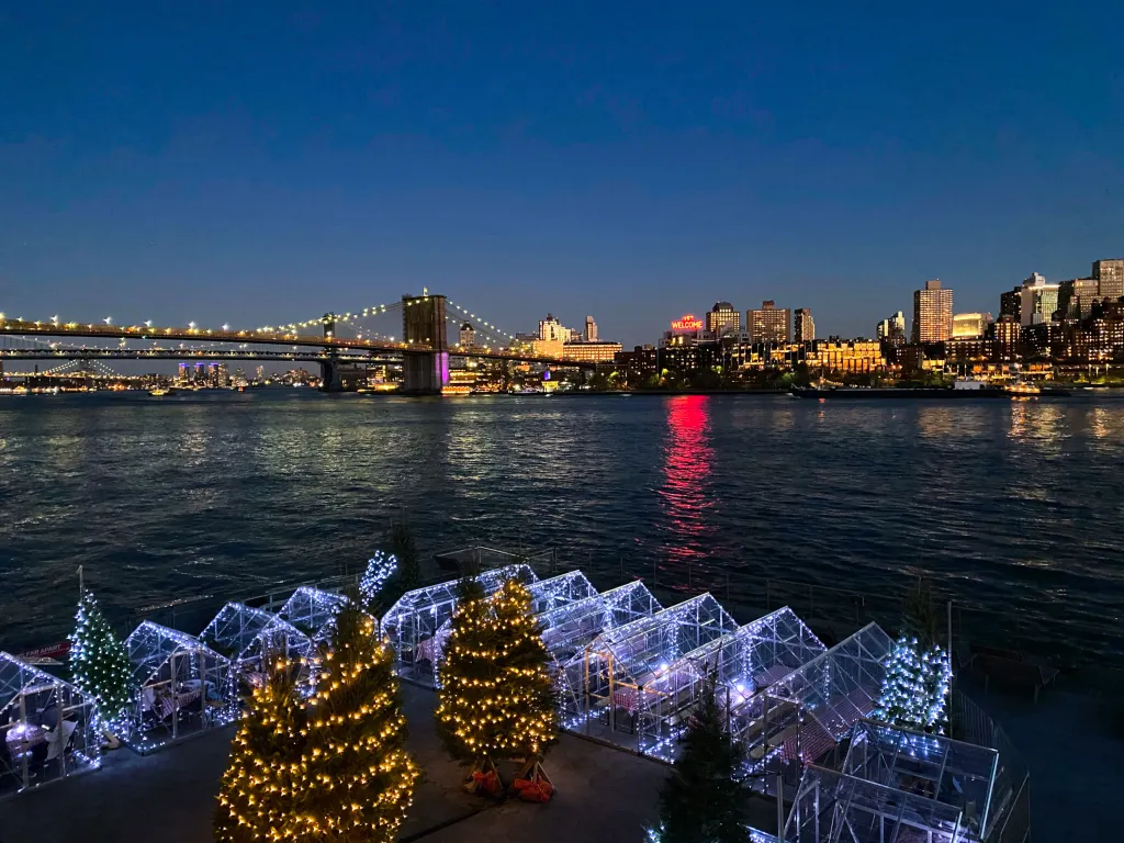 Waterfront restaurant with outdoor dining in illuminated individual greenhouses, Christmas trees, and a view of the Brooklyn Bridge at night.