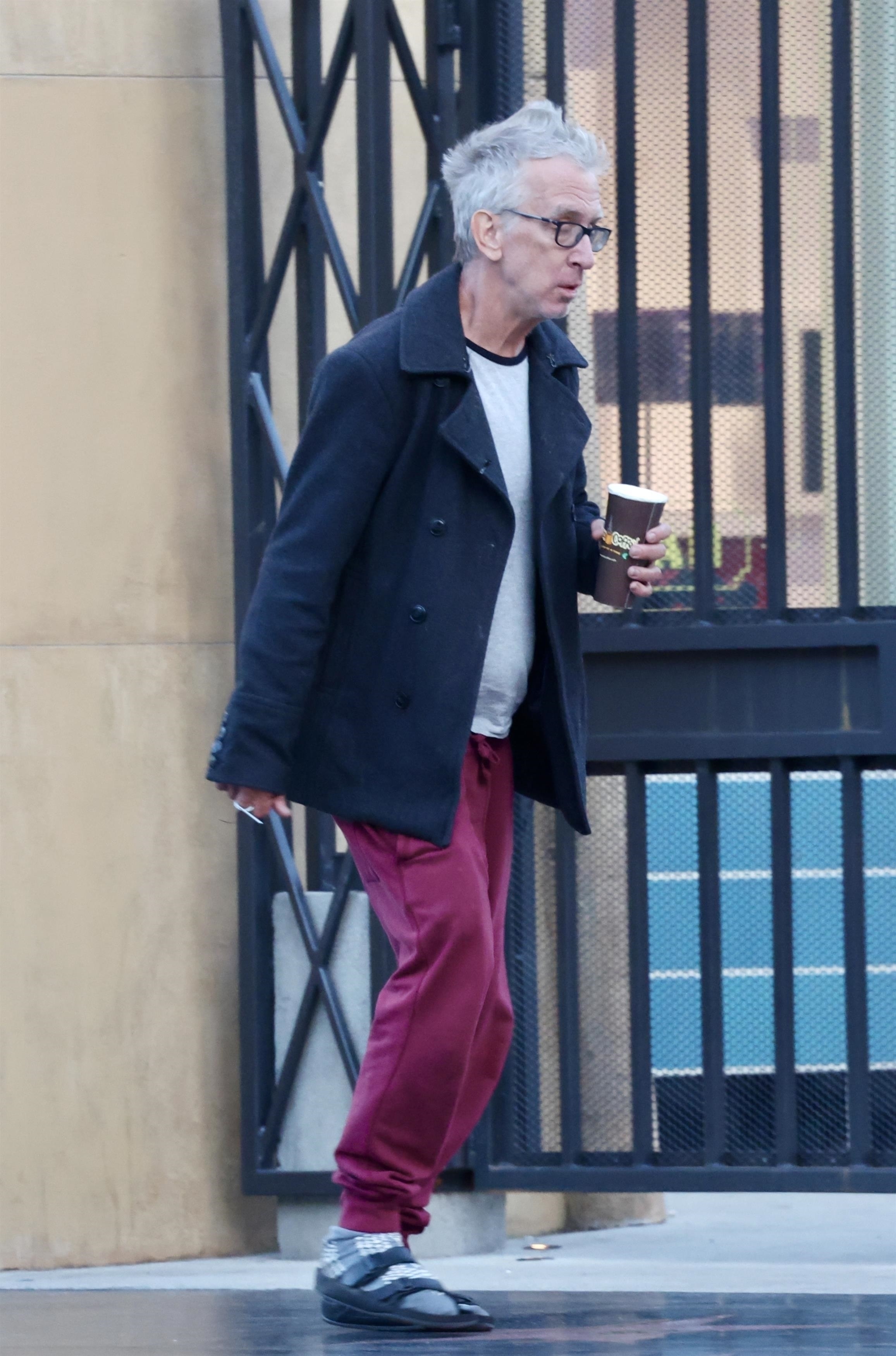 Andy Dick, wearing a black pea coat, gray t-shirt, maroon sweatpants, and sandals with socks, walking on a street while holding a coffee cup.
