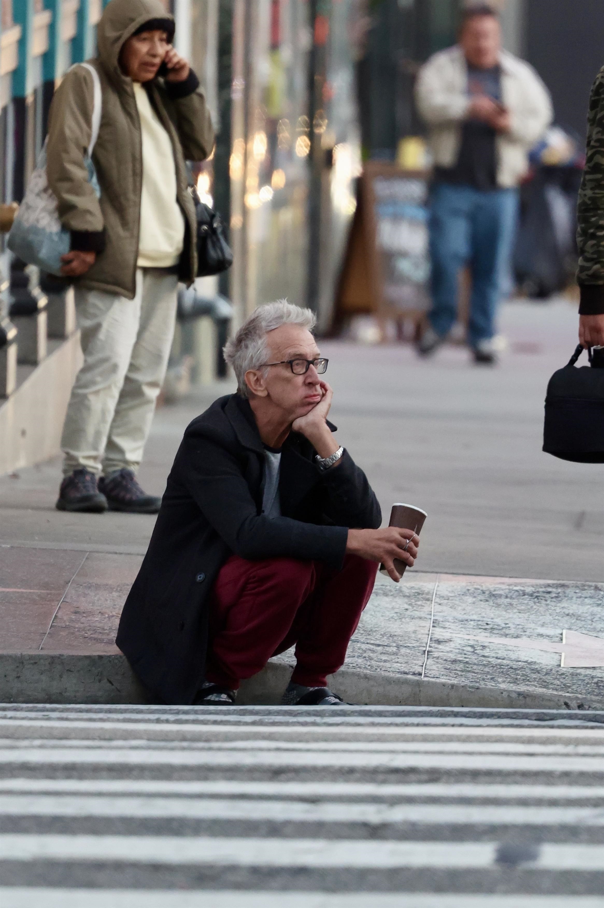 Andy Dick crouching on a street corner, holding a coffee cup, looking distraught.
